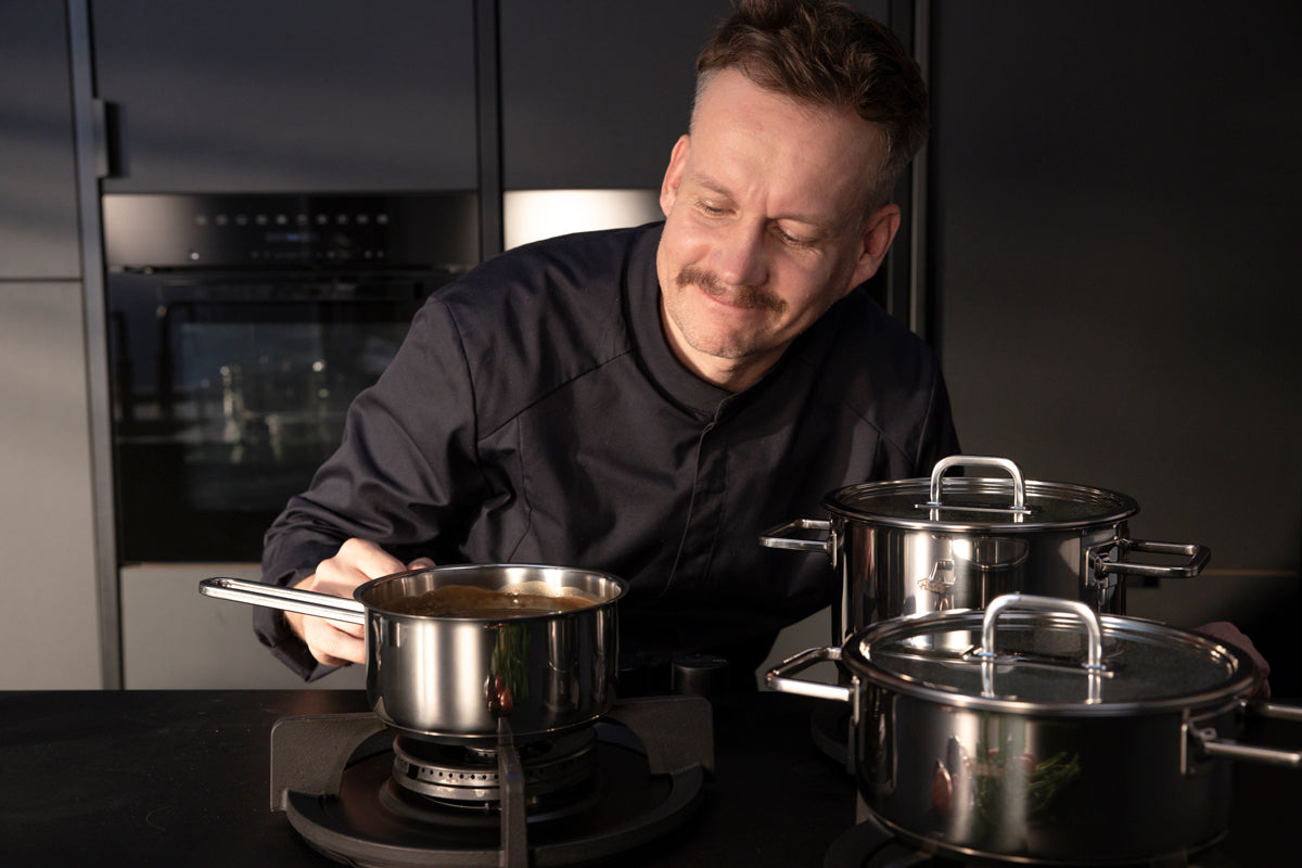 A chef in a modern kitchen enjoys a sauce simmering in a stainless steel pot, with Fissler Viseo® Pro pots nearby.
