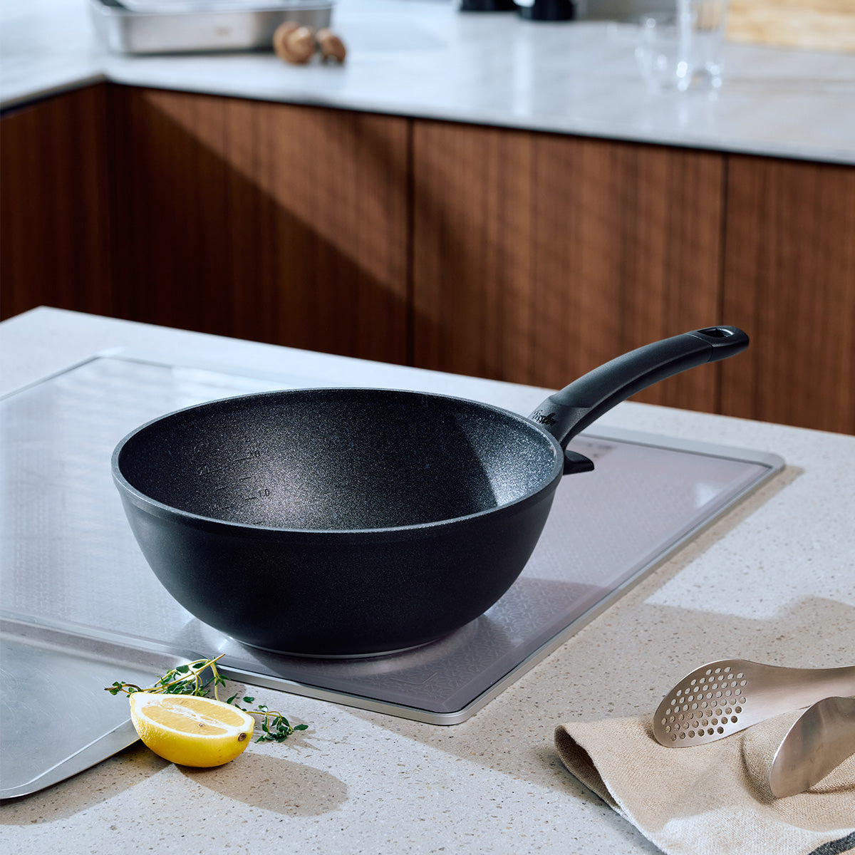 The Fissler Adamant® wok, with its sturdy handle and elegant black finish, sits stylishly on a marble countertop, with the warmth of wooden cabinets in the background. Made in Germany since 1845, it features durable, scratch-resistant non-stick coating.