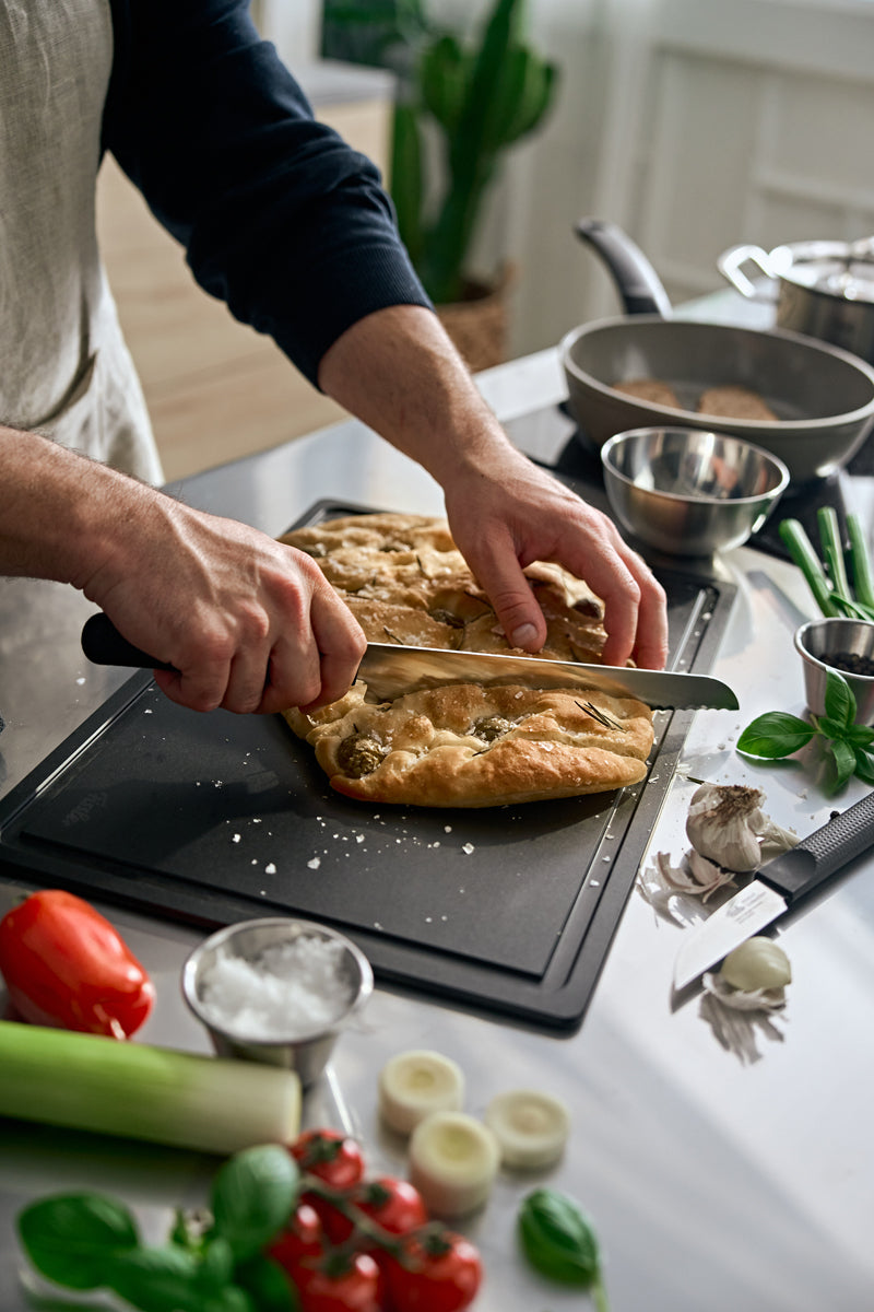 In a sunlit kitchen, focaccia is being sliced with the Fissler ProCut Bread Knife 21cm, surrounded by fresh ingredients and modern design.