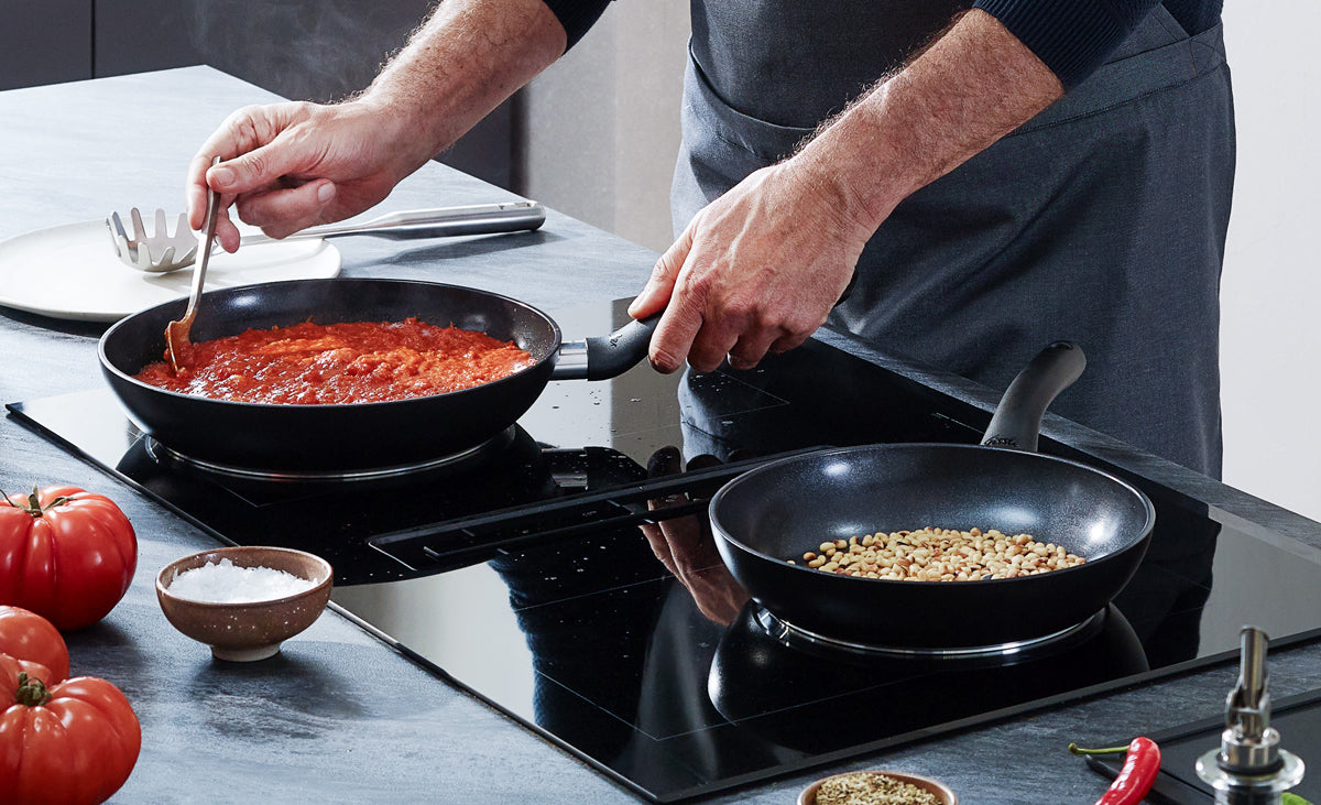 A chef prepares tomato sauce and roasts pine nuts with Fissler Essential® pans and stainless steel kitchen utensils on the counter.