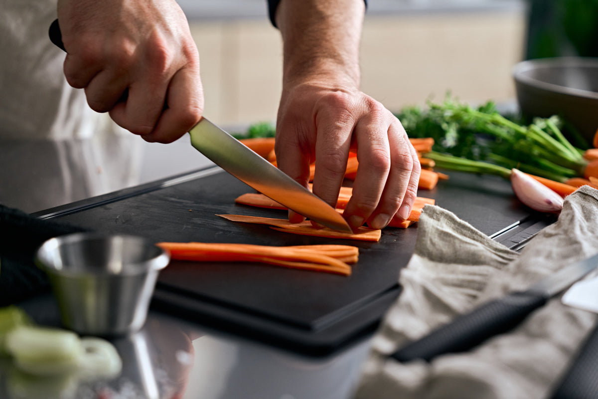 A person cuts carrots on a black cutting board with the Fissler ProCut Universal Knife 14cm; other Fissler knives and herbs nearby.