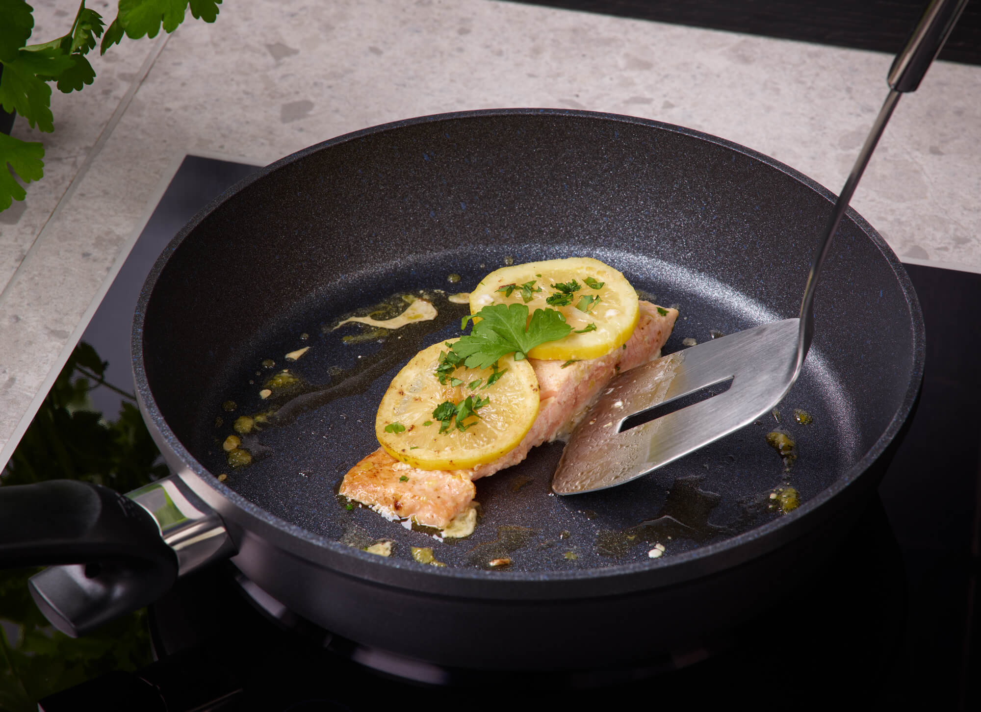 Lachsfilet mit Zitrone und Kräutern in einer Fissler Adamant® Comfort Pfanne, die mit einem Spatel angehoben wird; im Hintergrund frische Petersilie.