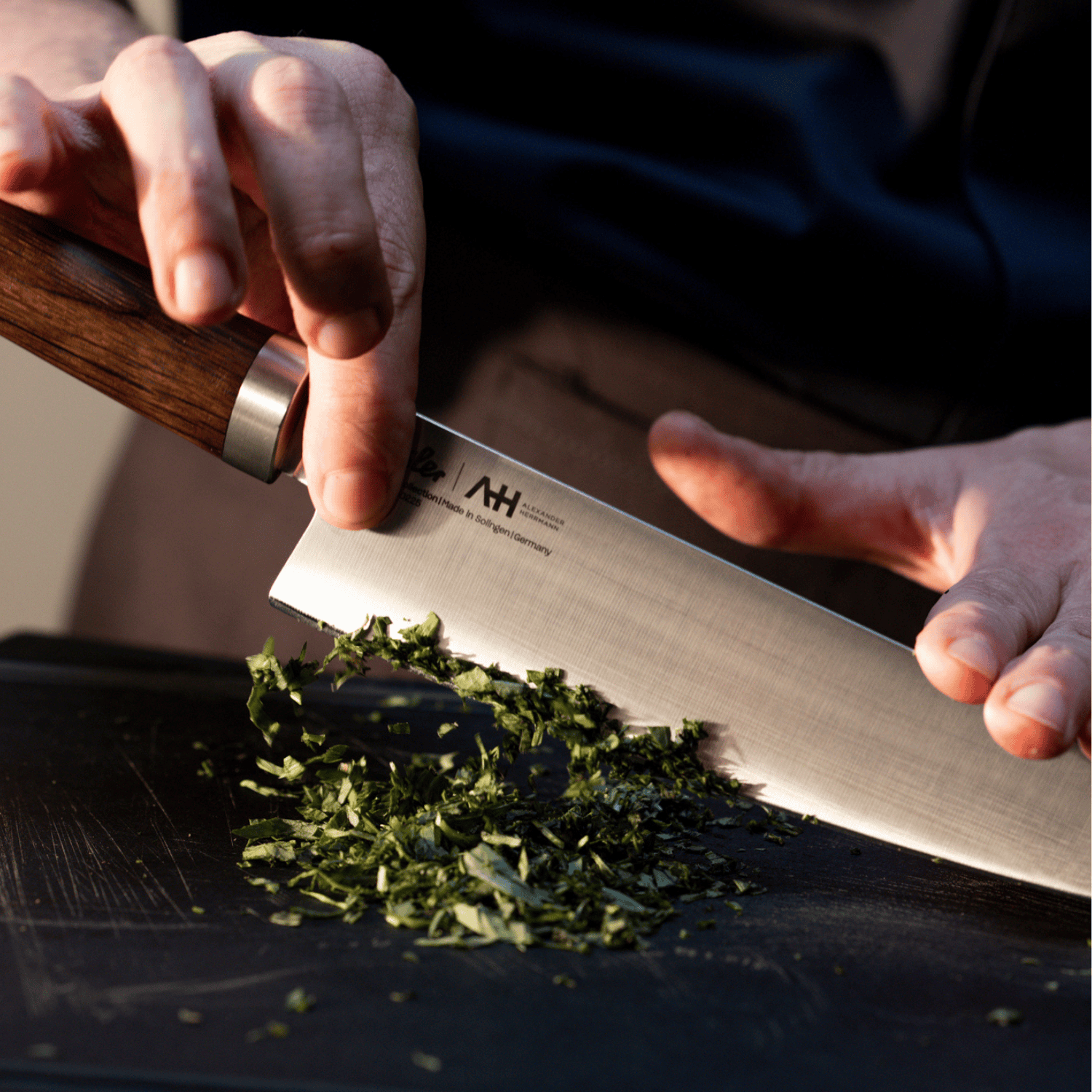 A person expertly chops fresh herbs with a Fissler precision chef's knife on a black cutting board, showcasing German craftsmanship.