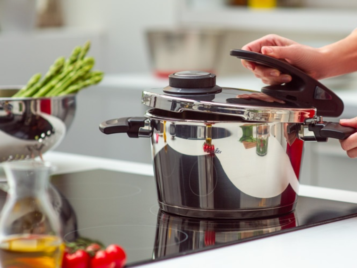 Eine Person öffnet einen Fissler Schnellkochtopf aus rostfreiem Stahl auf einem Herd; Spargel, Öl und Tomaten vervollständigen die hochwertige Küchenszene.