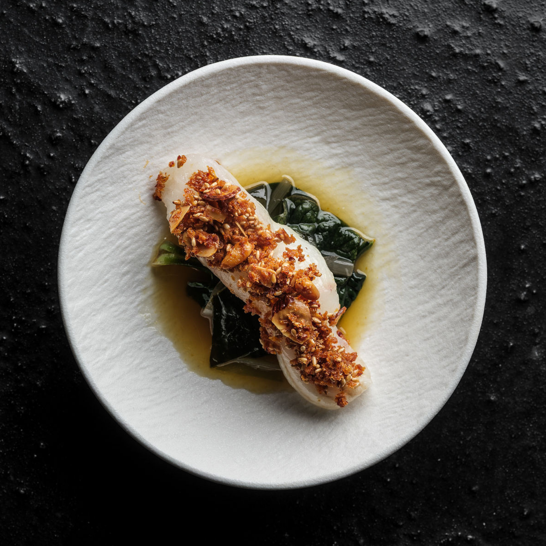 A perfectly cooked white fish fillet, finished with a golden crispy garnish, rests on a bed of fresh greens and savory broth in a pristine white bowl. Presented atop a textured black surface, this elegant dish showcases the art of precision cooking made possible with Fissler's expertly engineered cookware.