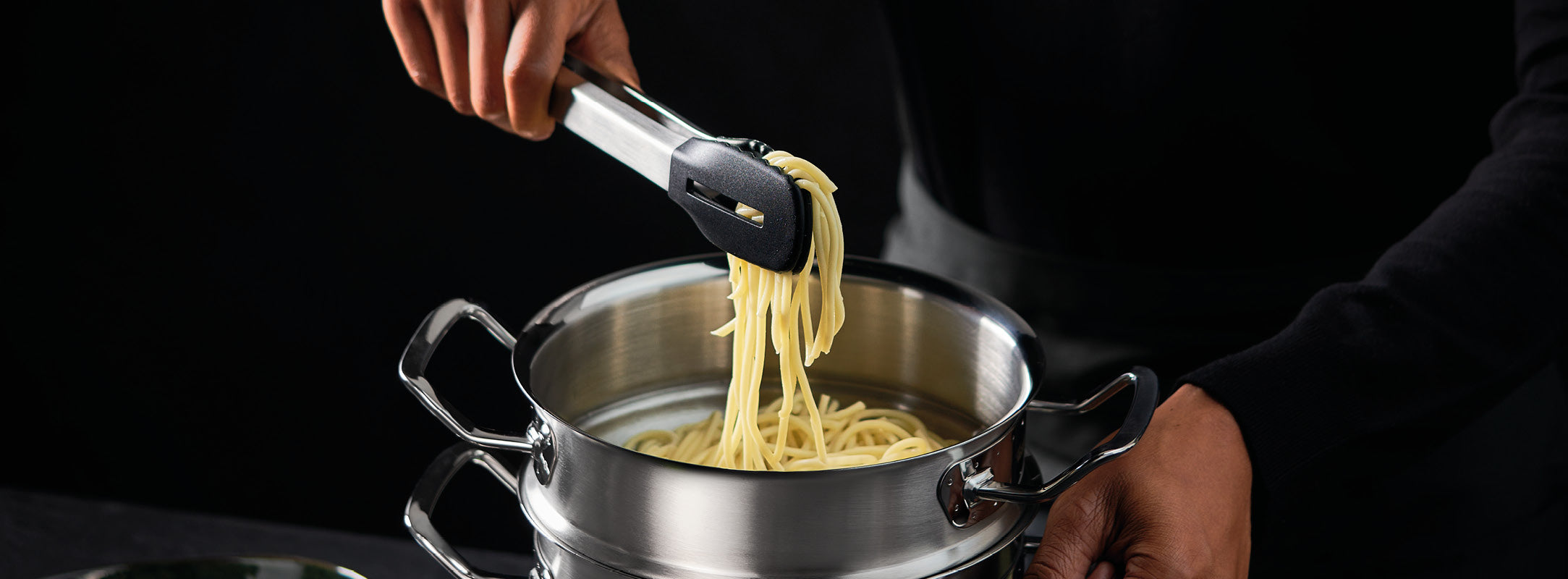 Eine Person hebt Spaghetti mit einer Zange aus einem Fissler-Edelstahltopf und unterstreicht damit dessen deutsche Qualität und zeitloses Design.