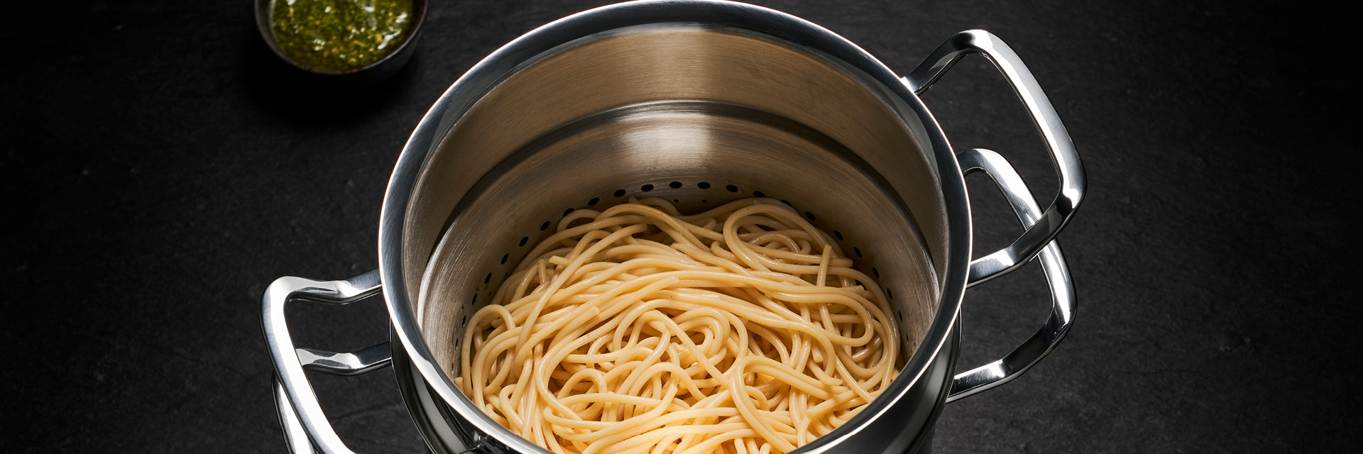 Ein Fissler-Edelstahltopf mit integriertem Sieb enthält frisch gekochte Spaghetti, die mit grünem Pesto auf einer dunklen Oberfläche kombiniert sind.