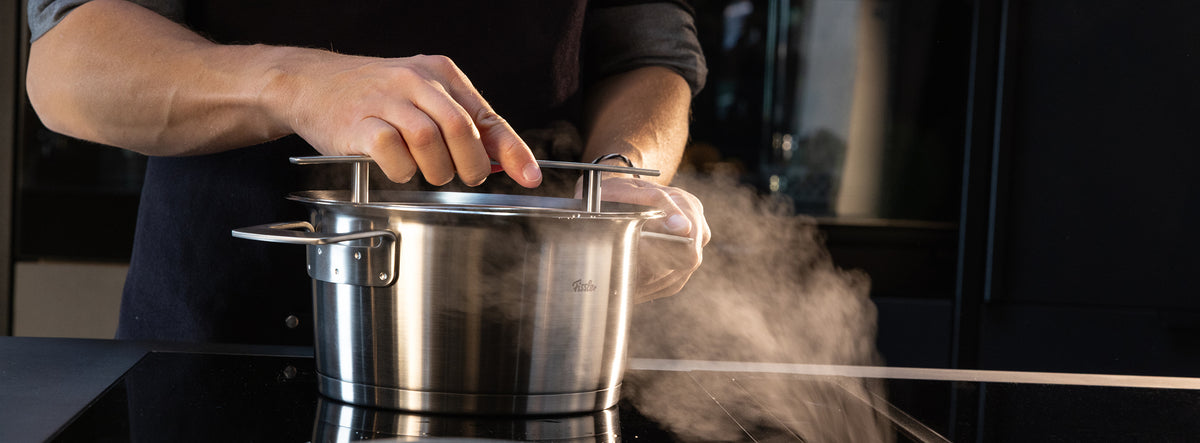 Eine Person hebt den Deckel eines Fissler-Edelstahltopfes auf einer Herdplatte an und zeigt dabei, wie der Dampf aufsteigt.