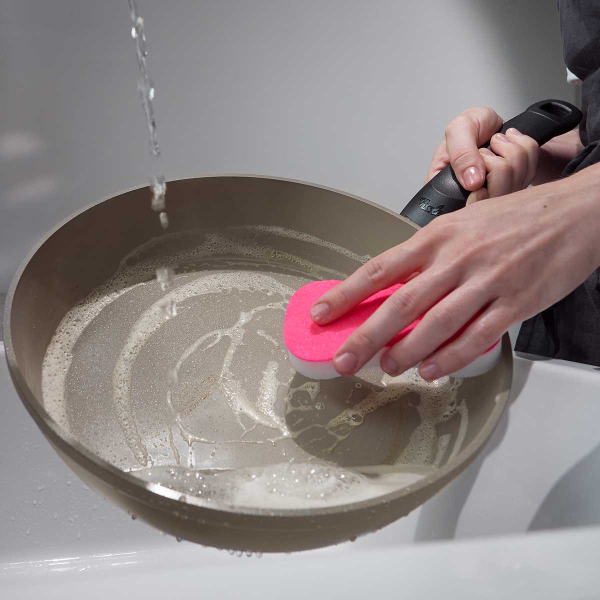 Eine Person reinigt ein Fissler Ceratal® Comfort Pfannenset (24 cm & 28 cm) mit einem rosafarbenen Schwamm unter fließendem Wasser und demonstriert damit deutsche Qualität.
