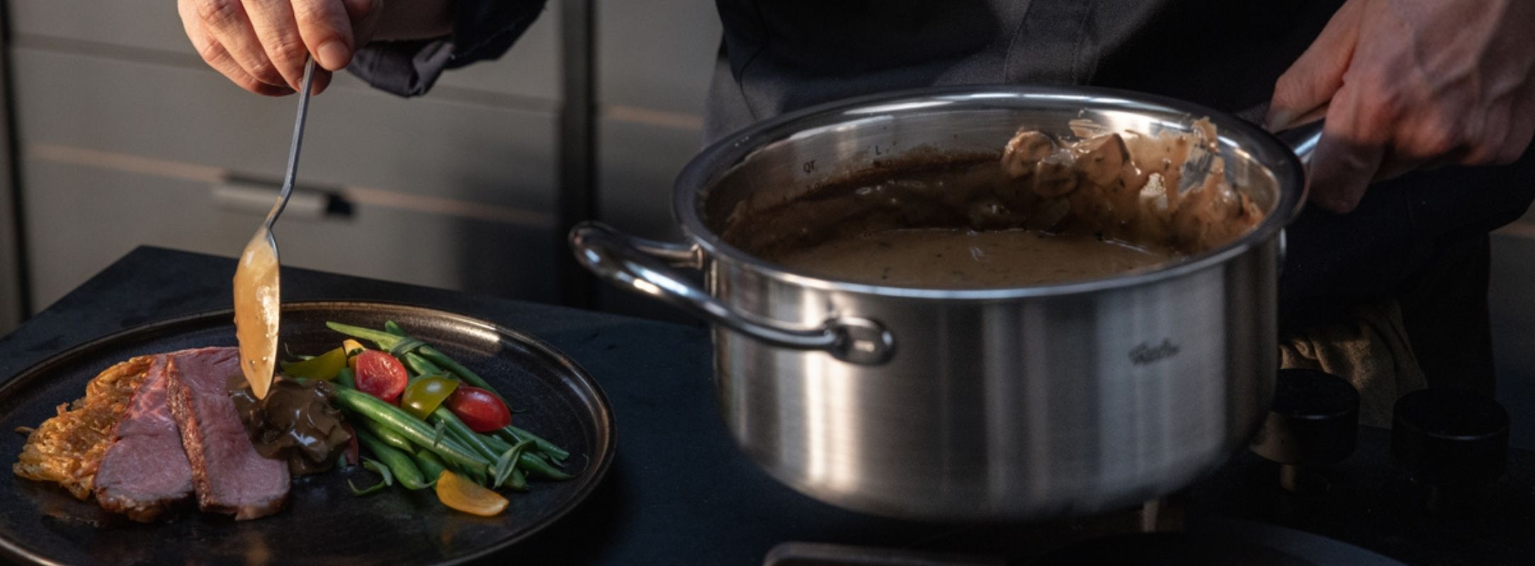 A home cook serves sauce from a Fissler stainless steel pot onto a plate with sliced meat and vegetables in a modern kitchen.