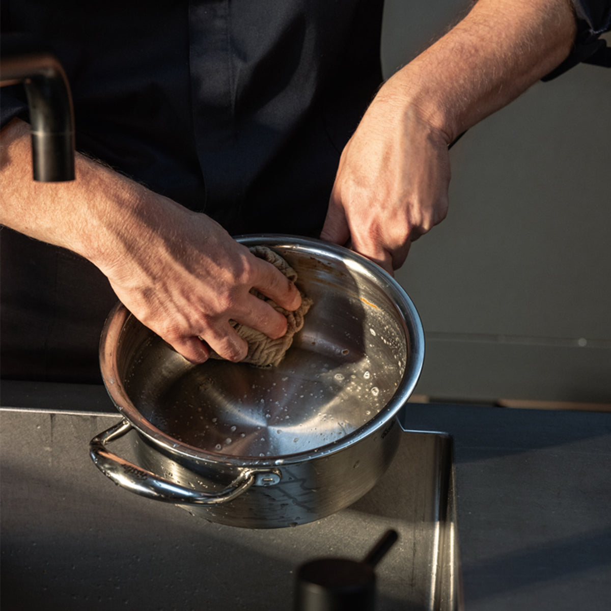 A person carefully cleans the Fissler M5 Pro-Ply® Stielkasserolle mit Metalldeckel—crafted in Germany from recycled stainless steel—using a sponge at the kitchen sink, washing away soap and residue. Engineered for durability and effortless care, this dishwasher-safe saucepan with induction compatibility exemplifies Fissler’s commitment to innovation, sustainability, and timeless design.