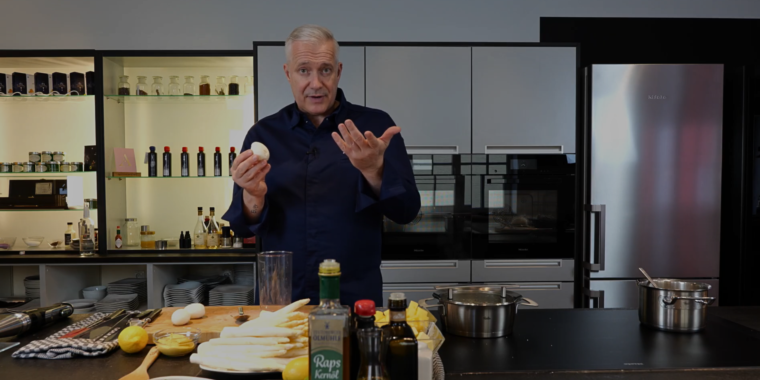 Ein Mann in einer modernen Küche hält ein geschältes Ei von Fissler Kochgeschirr, mit Eiern, Spargel, Flaschen auf einem Holzbrett vor ihm.