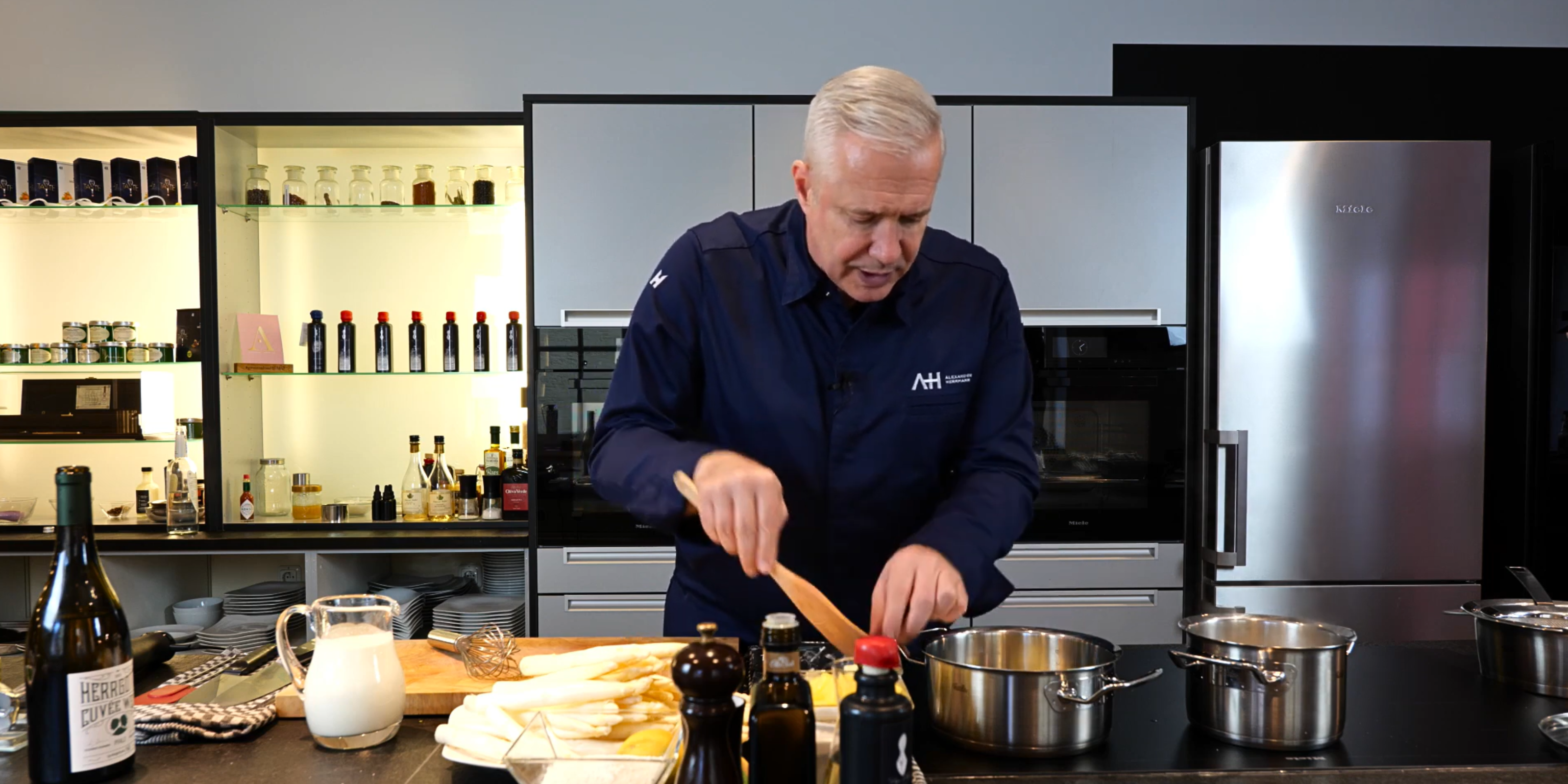Ein Mann in einer marineblauen Kochjacke benutzt Fissler-Edelstahlkochgeschirr in einer eleganten Küche, frische Zutaten liegen auf dem Tresen daneben.