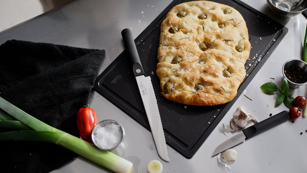 Eine rechteckige Focaccia auf einem Brett neben dem Fissler ProCut Brotmesser 21cm, frische Produkte und Küchenakzente auf einer grauen Fläche.
