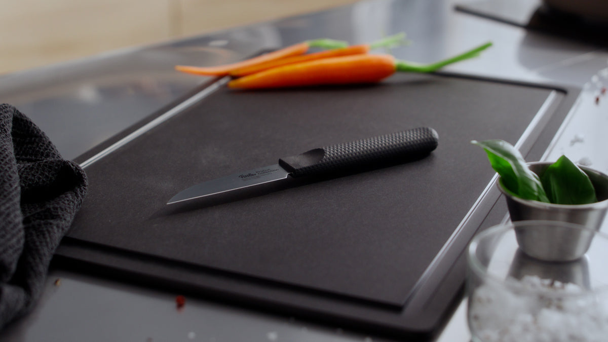 Fissler ProCut 9cm Vegetable Knife on a black board with carrots, herbs in a steel bowl, and a towel. Precision meets timeless German design.