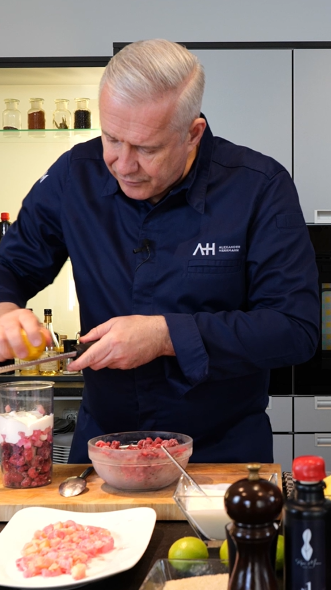 Ein Koch reibt Zitronenschale über Fleisch und Zwiebeln auf einer hölzernen Arbeitsplatte und verwendet dazu hochwertiges Fissler-Kochgeschirr in einer eleganten, modernen Küche.