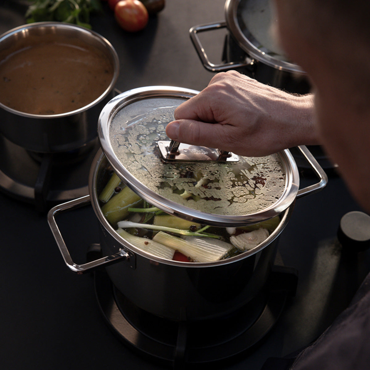 A hand lifts the Fissler Viseo® Pro 20 cm glass lid from a Viseo Pro pot; another Fissler pot with sauce simmers next to fresh vegetables.