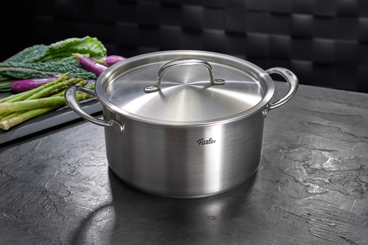 Das Fissler M5 Pro-Ply® 4-teilige Topfset mit Metalldeckeln, gefertigt aus recyceltem Edelstahl “Made in Germany”, präsentiert sich mit robusten, ergonomischen Griffen auf einer dunklen Arbeitsplatte. Im Hintergrund sorgen frischer Spargel und Blattgemüse für Kochinspiration. Dieses hochwertige Set verkörpert Fisslers Tradition aus Präzision, Langlebigkeit und innovativem Design – optimal für Induktionsherde sowie vielseitiges Kochen.