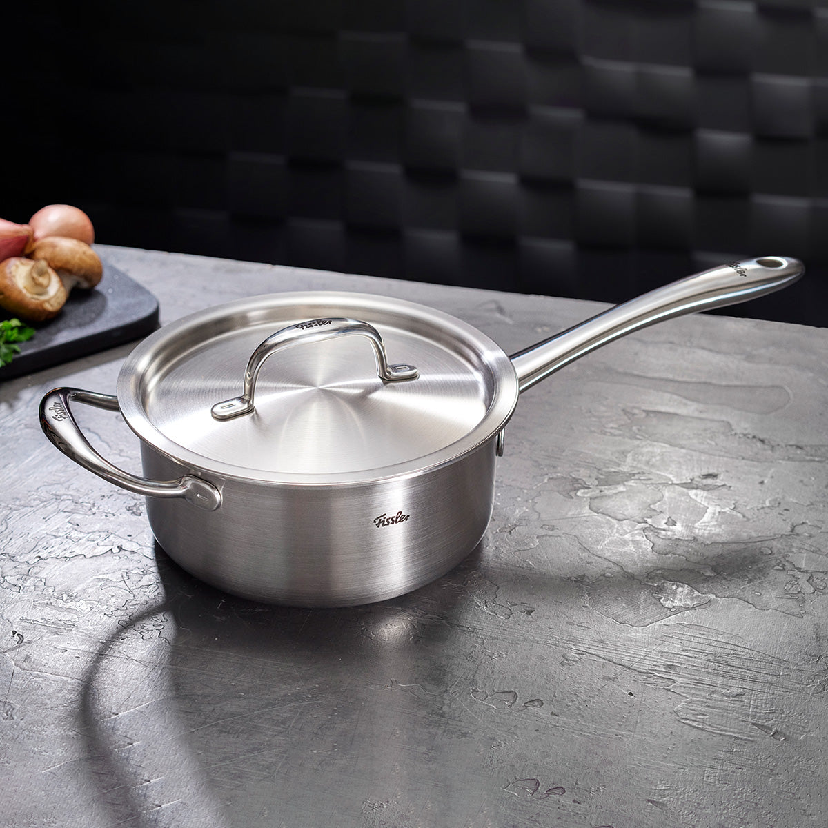 Fissler M5 Pro-Ply stainless steel saucepan with lid on a gray surface, with a cutting board featuring mushrooms and onions in the background.