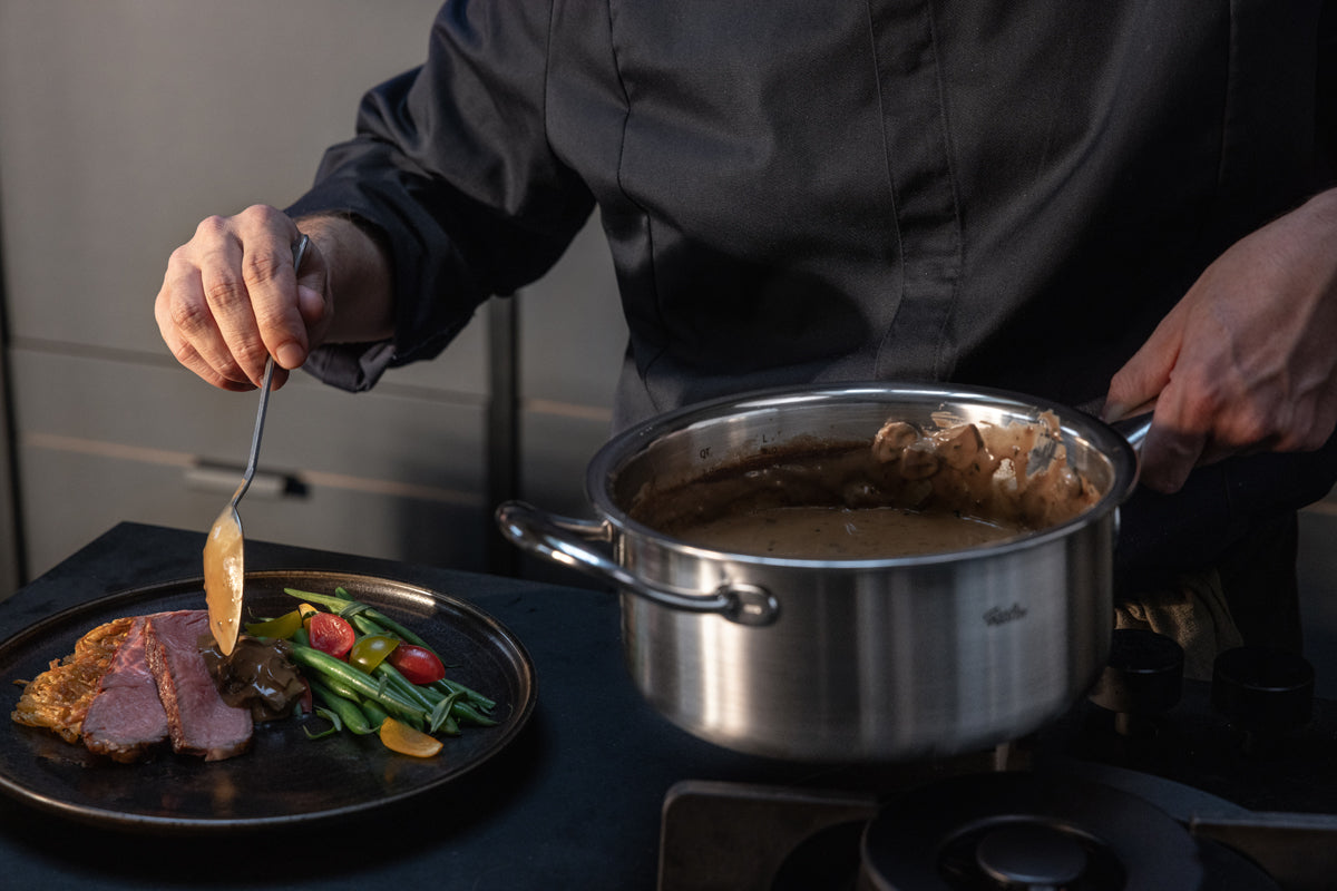 A chef in dark attire pours sauce from a Fissler stainless steel pot onto sliced meat beside vibrant vegetables on a black plate, demonstrating the precision and quality of the Fissler M5 Pro-Ply® 3-piece set with metal lid in a modern kitchen. The scene highlights Fissler’s heritage of German engineering, durable materials, and innovation for timeless cooking performance.