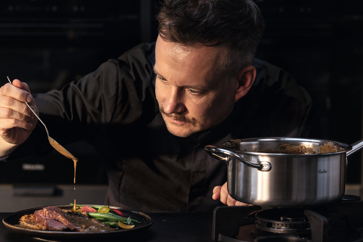 A chef in a black jacket expertly drizzles sauce over a plated dish of vegetables and meat, using a spoon crafted from recycled stainless steel. Beside the chef on the stove is the Fissler M5 Pro-Ply® Stielkasserolle mit Metalldeckel—an induction-compatible saucepan renowned for its precision engineering, exceptional durability, and timeless German design.