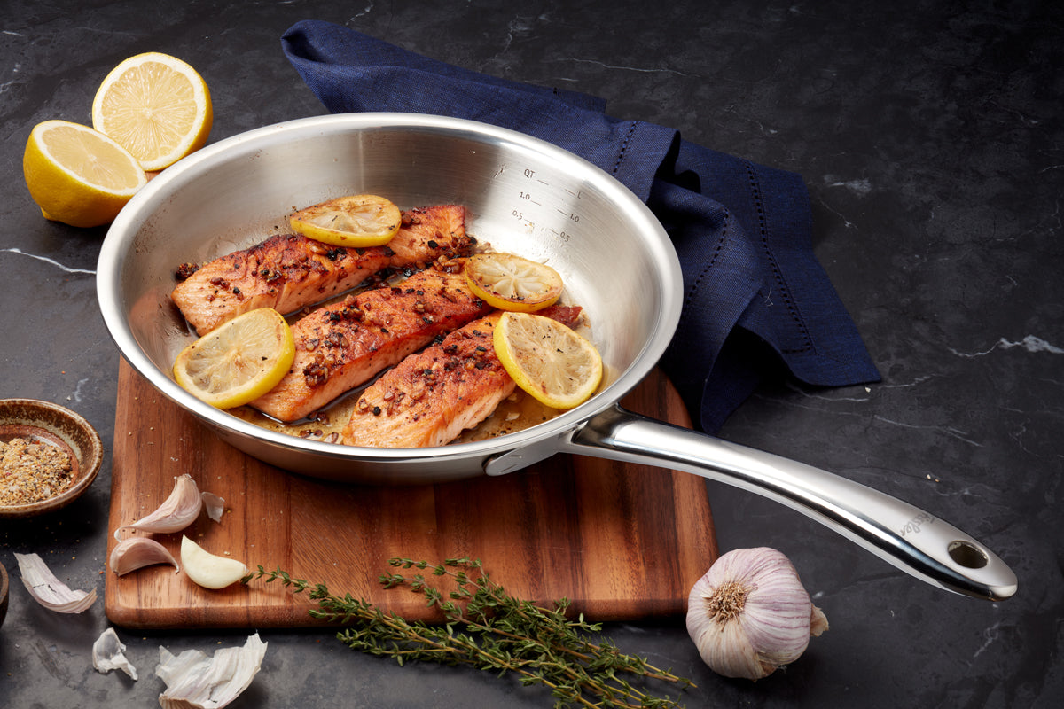 Three perfectly cooked salmon filets with lemon slices are presented in a Fissler M5 Pro-Ply® 3-piece pan with metal lid, displayed on a wooden board and accented by fresh herbs, garlic cloves, lemon halves, and a small spice bowl. A dark napkin completes the elegant kitchen setting. This scene highlights Fissler’s tradition of precision German engineering and timeless design—our premium stainless steel cookware brings durability and innovation to every modern kitchen.