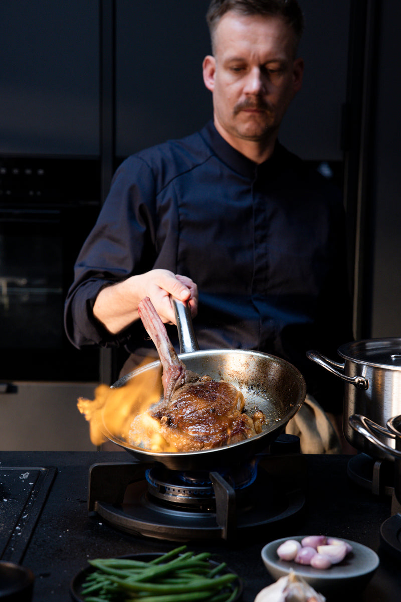 A chef expertly sears a steak in a Fissler M5 Pro Ply® pan; flames rise, showcasing German quality and innovation.