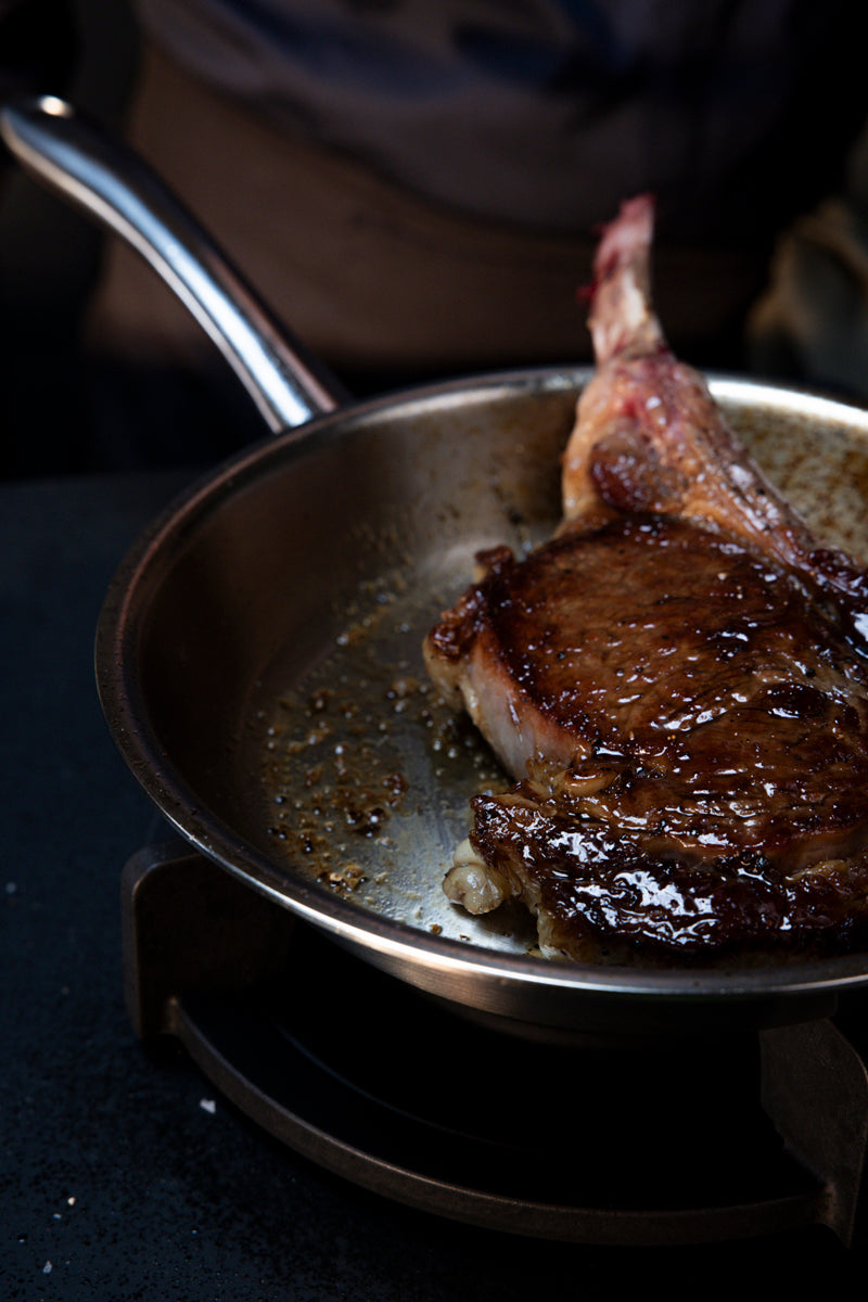 Ein Steak mit Knochen lässt sich im Fissler M5 Pro-Ply® 3-tlg. Set mit Metalldeckel perfekt anbraten, wobei der reichhaltige Bratensaft und die karamellisierten Stückchen die hervorragende Verarbeitung der glänzenden Edelstahlpfanne unterstreichen. Dezente Beleuchtung unterstreicht die zeitlose deutsche Ingenieurskunst und Langlebigkeit, die das Fissler-Kochgeschirr auszeichnen – induktionsgeeignet für müheloses, vielseitiges Kochen.