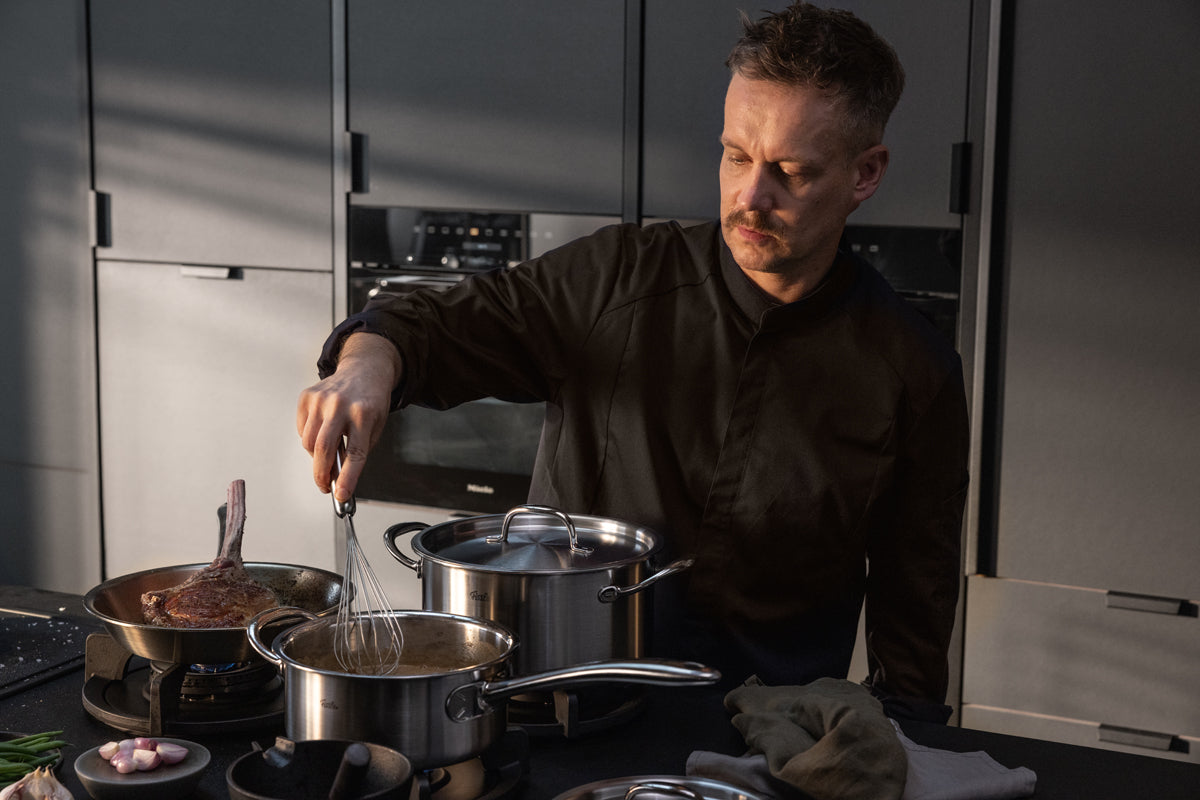 A man in a black shirt expertly whisks food in a premium Fissler stainless steel pot on the stove, alongside a pan from the Fissler M5 Pro-Ply® 4-piece set with metal lid made from recycled stainless steel, surrounded by other high-quality kitchen items in a sleek, modern kitchen—demonstrating Fissler’s heritage of German precision engineering and timeless design.