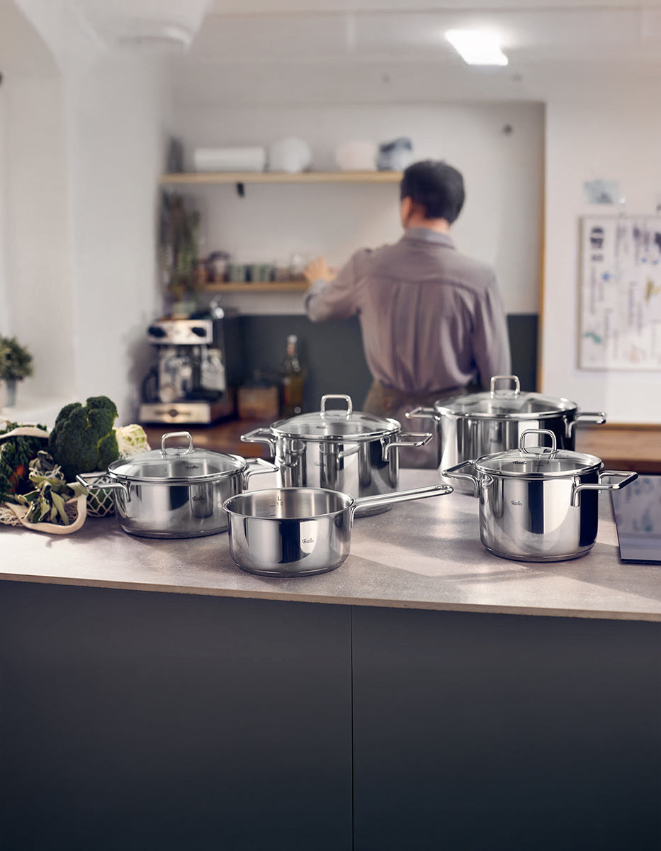 Eine Küche mit dem 5-teiligen Fissler LumiTherm Topfset aus Edelstahl, frischem Gemüse und einer Person mit Regalen und Kaffeemaschine.