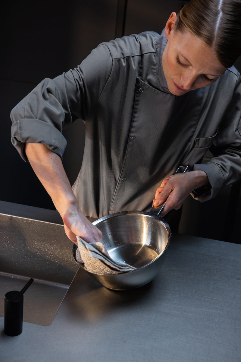 A chef in gray dries the 6-piece Fissler Original-Profi Collection® stainless steel set with glass lids on an elegant counter.