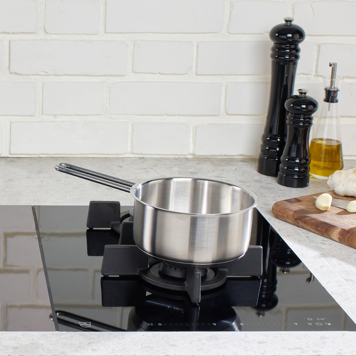 A Fissler Pure Collection Stielkasserolle, crafted from premium stainless steel in Germany, sits centered on a sleek black gas stove. Beside it are classic black pepper and salt grinders, a bottle of oil, and a wooden cutting board with fresh garlic cloves—each piece echoing professional kitchens. The white brick wall in the background highlights the timeless design and superior quality that define Fissler’s heritage cookware collection since 1845.