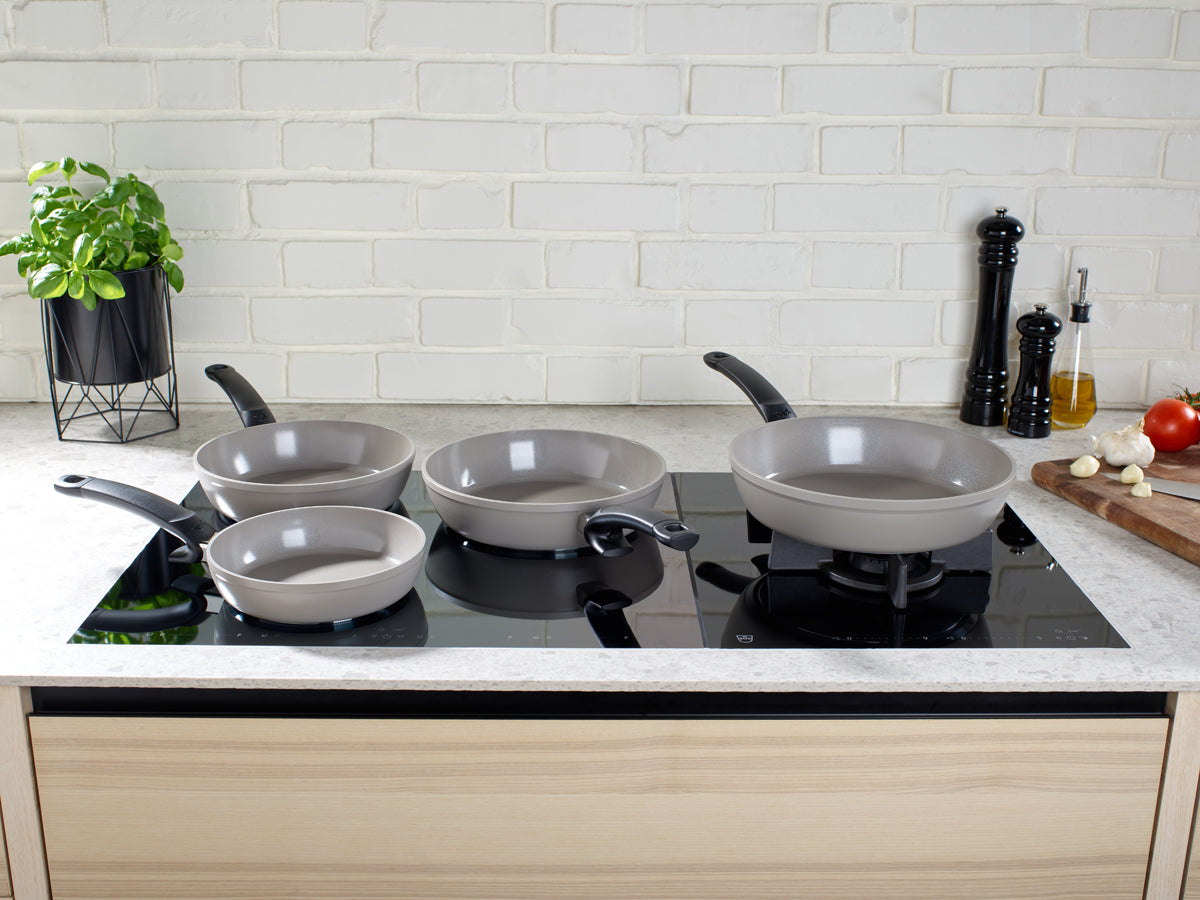 Zwei graue Fissler Ceratal Comfort Pfannen (24cm & 28cm) mit Keramikbeschichtung auf schwarzem Induktionskochfeld, umgeben von frischen Kräutern.