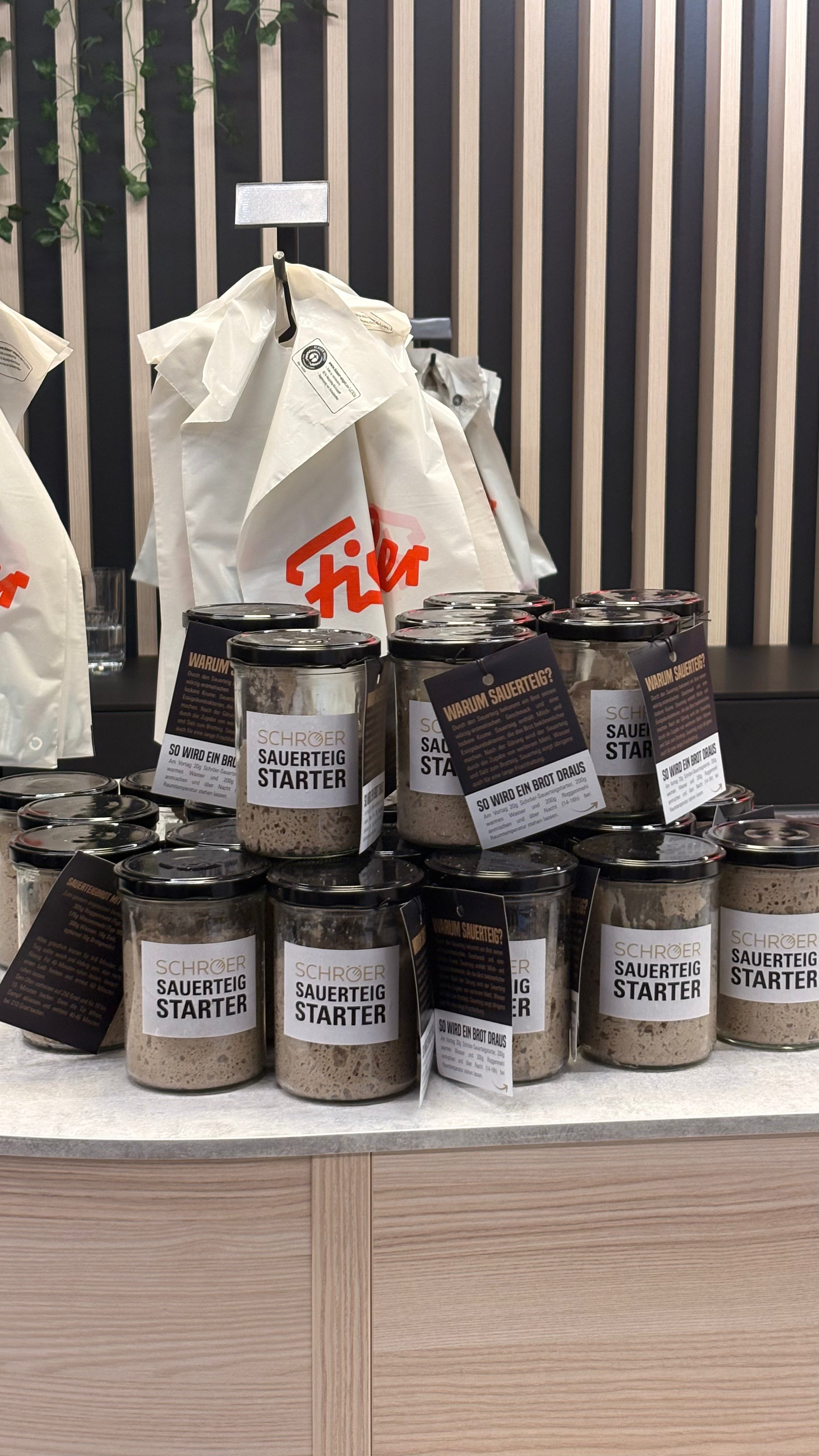 Jars of premium Fissler sourdough starter are neatly stacked on a countertop, highlighting our tradition of quality and innovation. In the background, durable tote bags with bold red Fissler logos hang against a sleek black and wood-paneled wall, evoking timeless German design and craftsmanship.