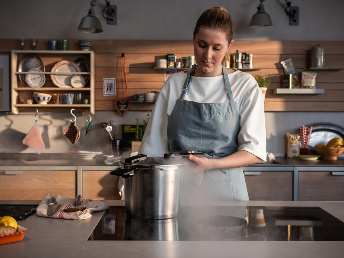 A woman in a blue apron stands in a sunlit kitchen, carefully releasing steam from her Fissler Vitaquick® Premium 2-piece pressure cooker set with SKT Book on the stove. Designed and engineered in Germany since 1845, our cookware exemplifies precision, durability, and timeless innovation. Shelves stocked with dishes and kitchen essentials provide a warm backdrop that perfectly complements the elegant stainless steel construction of this premium Made in Germany Schnellkochtopf Set.