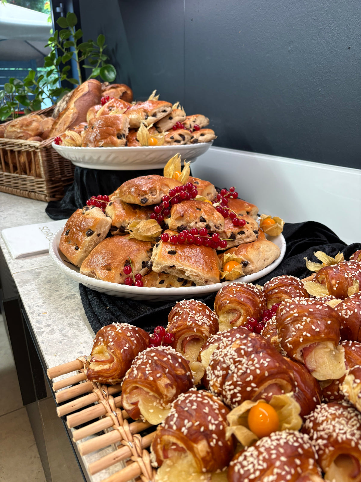 Assorted pastries such as chocolate chip scones and sesame-topped croissants, garnished with berries and cape gooseberries, elegantly arranged on platters and trays atop a countertop showcasing Fissler’s signature stainless steel serveware—embodying timeless German craftsmanship and kitchen innovation.