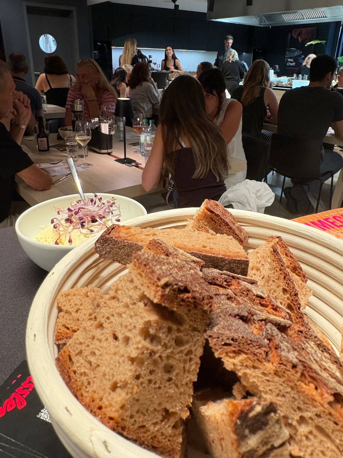 A close-up of a basket filled with sliced bread on a modern kitchen table, accompanied by a fresh salad in a Fissler stainless steel bowl. In the background, people dine and interact in an inviting kitchen space featuring premium Fissler cookware—showcasing precision engineering and timeless German design.