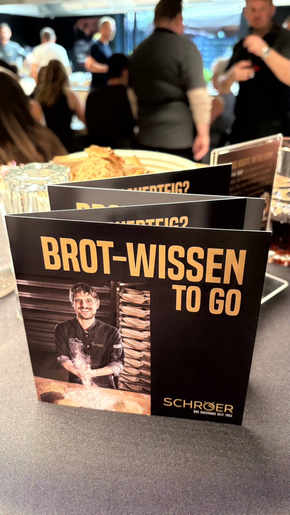 A brochure titled Brot-Wissen To Go with a smiling baker on the cover is elegantly displayed on a table at a vibrant event, surrounded by other brochures, sleek glassware, and attendees in the background. The scene reflects Fissler’s commitment to German heritage and innovation in culinary craftsmanship—an inviting atmosphere where high-quality cookware such as stainless steel pots, pans, pressure cookers, and kitchen accessories seamlessly complement the setting.