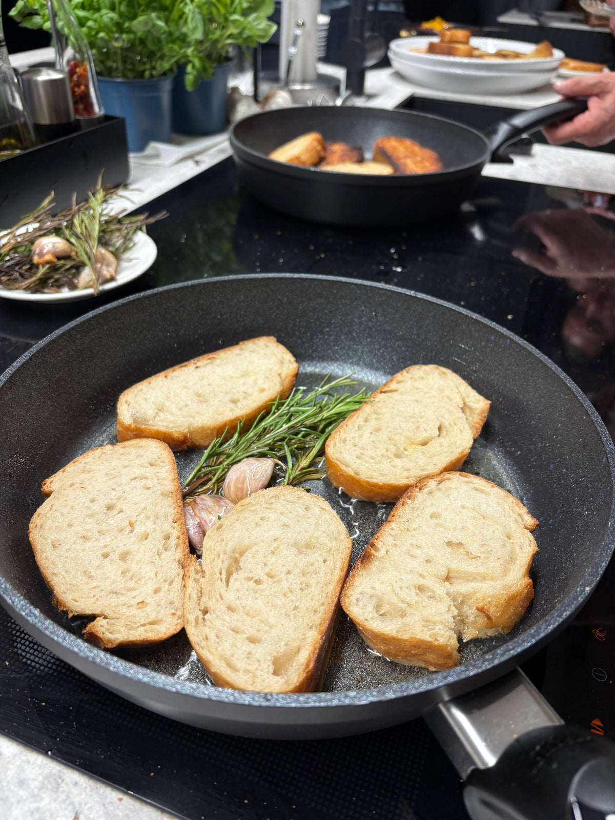 Five slices of bread are perfectly toasting with fresh rosemary and garlic in a precision-engineered Fissler pan, showcasing even browning and superior heat distribution. In the background, another high-quality Fissler pan with crisping bread and a plate of aromatic herbs and garlic rest on a modern kitchen counter, reflecting the brand’s commitment to innovative design, durability, and German craftsmanship.