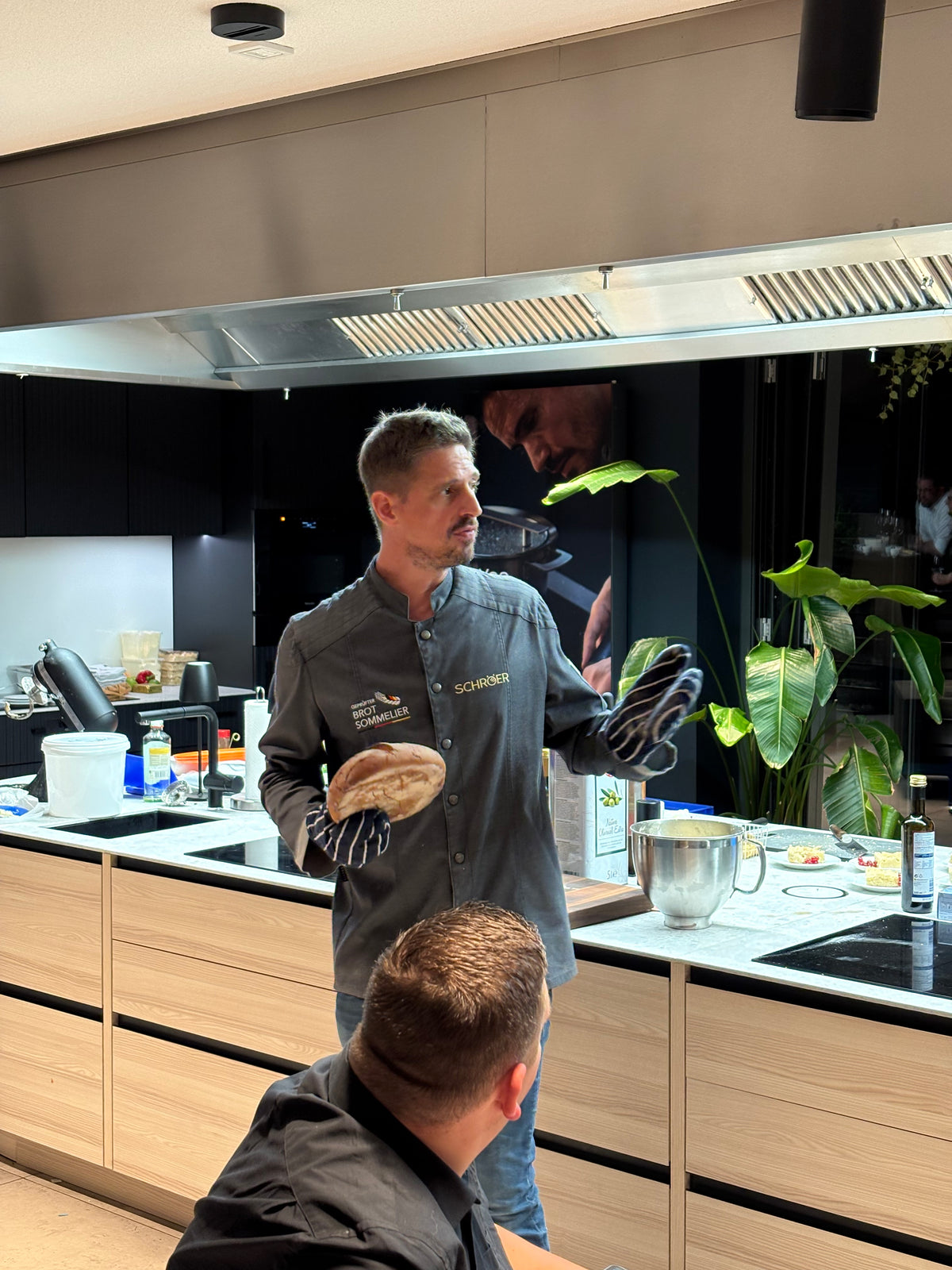 A chef in a gray uniform holds a loaf of bread and an oven mitt in a modern kitchen, engaging with another person. On the counter behind him, premium Fissler stainless steel cookware, kitchen accessories, and fresh ingredients are displayed alongside plants, reflecting the brand’s commitment to precision engineering, durability, and timeless German design.