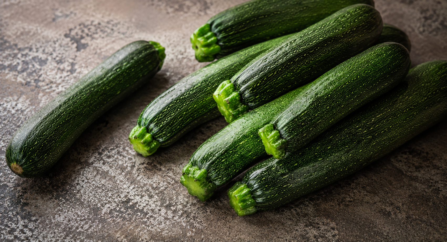 Eine Auswahl leuchtend grüner Zucchini liegt auf einer braun gesprenkelten Arbeitsplatte und unterstreicht, wie ideal sich diese frischen Zutaten für die Zubereitung in Fisslers präzise gefertigten Edelstahl-Kochgeschirren eignen. Eine Zucchini liegt etwas abseits – ein Beispiel für die Liebe zum Detail und die Qualität, die unsere in Deutschland hergestellten Küchenaccessoires und Kochutensilien seit 1845 auszeichnen.
