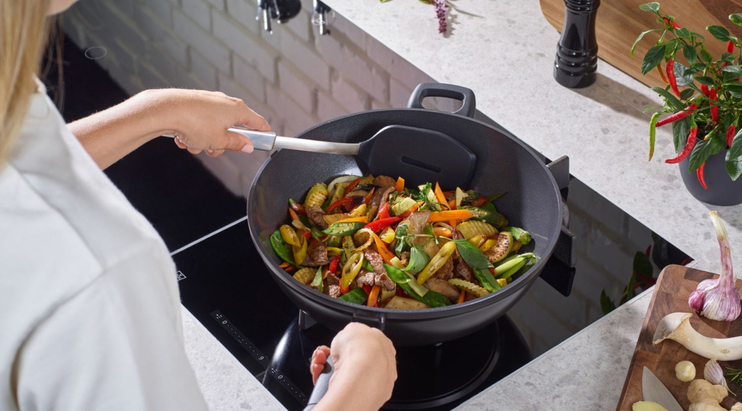 Ein Koch bereitet in einem schwarzen Premium-Wok von Fissler auf einem modernen Herd ein lebhaftes Wokgericht zu, wobei er frische Zutaten und Werkzeuge zur Hand hat.