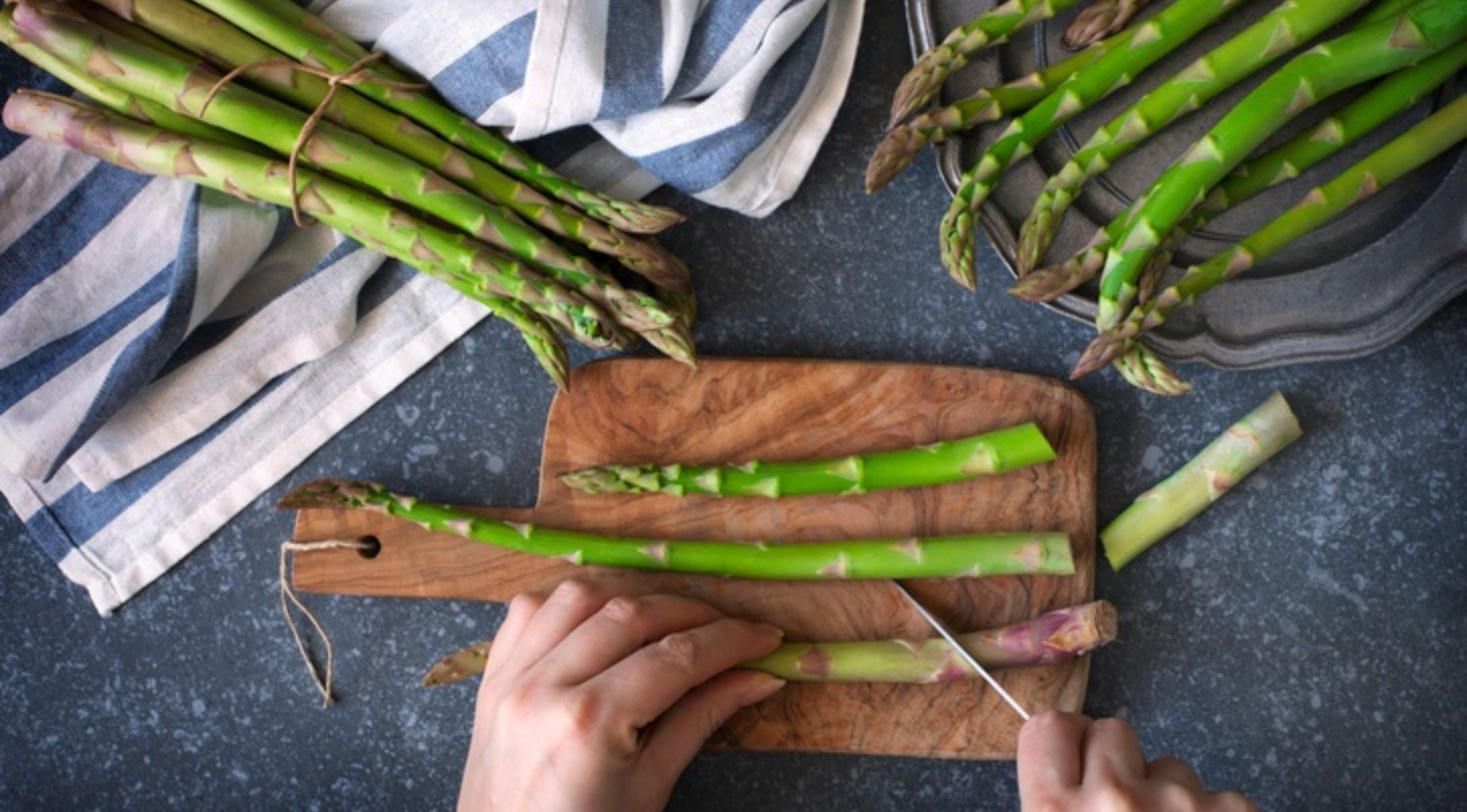 Eine Person schneidet grünen Spargel auf einem Holzbrett, während Fissler-Kochgeschirr und Küchentücher auf einem schlichten grauen Tresen stehen.