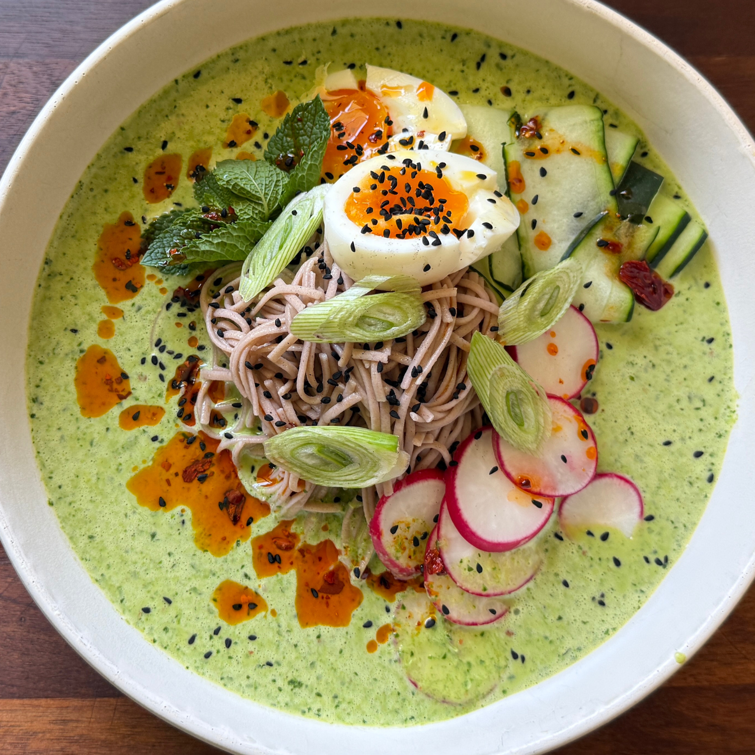 Eine wunderschön angerichtete Schüssel mit grüner Brühe, Soba-Nudeln, Radieschenscheiben, Gurke, Frühlingszwiebeln, frischer Minze, einem halbierten weichgekochten Ei, schwarzem Sesam und Chiliöl. Das optisch beeindruckende Gericht wird so präsentiert, dass die Liebe zum Detail deutlich wird – zubereitet mit Küchenutensilien wie dem präzisionsgefertigten Kochgeschirr von Fissler, das optimale Garergebnisse und zeitloses Design vereint.