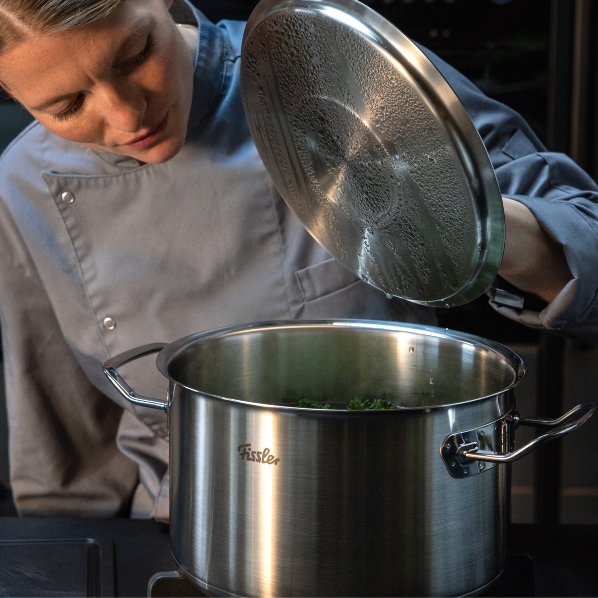 A chef in a gray uniform lifts the lid of a Fissler stainless steel pot on the stove, revealing perfectly controlled steam and condensation—a testament to the precision engineering and timeless design of Fissler cookware, made in Germany.