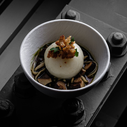 A white bowl featuring a perfectly poached egg resting on sliced mushrooms in a rich, dark broth, garnished with golden crispy crumbs and fresh herbs. The dish is presented on a robust metal surface with prominent bolts—a subtle nod to Fissler’s precision-engineered German cookware that inspires culinary excellence and durability.