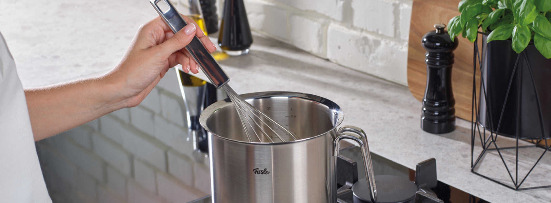 Eine Person rührt Zutaten in einem hochwertigen Fissler-Edelstahltopf auf dem Herd um, während in der Nähe hochwertige Küchengeräte elegant angeordnet sind.