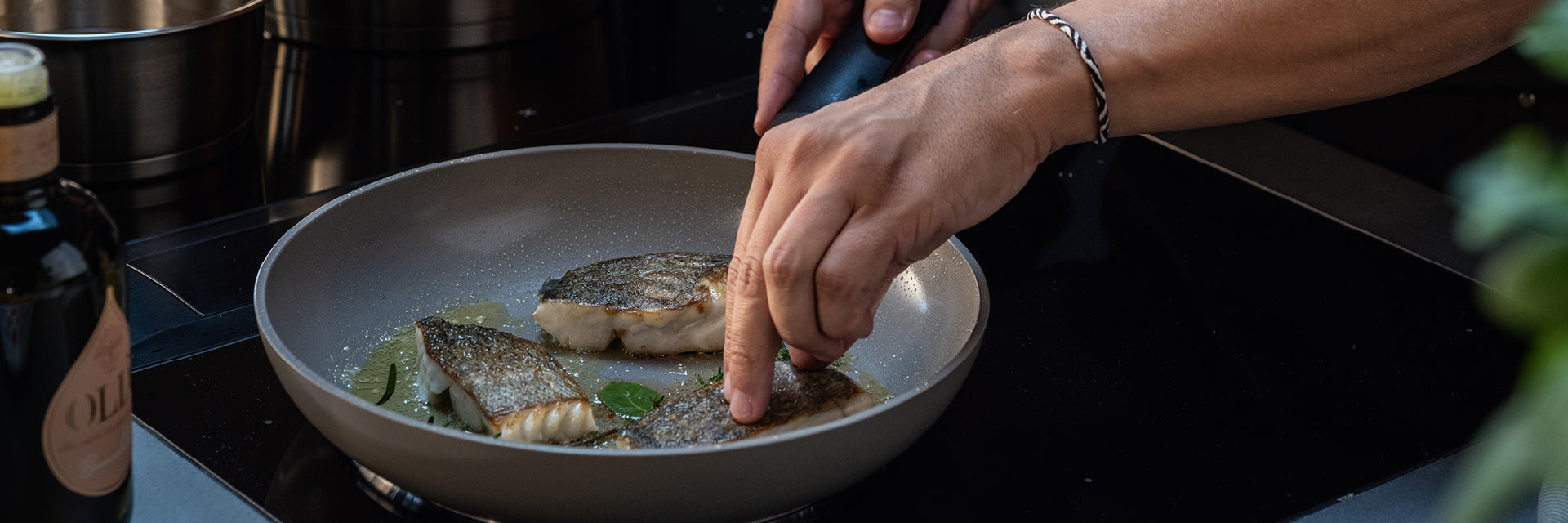 Eine Person wendet Fischfilets in einer Fissler-Pfanne auf dem Herd mit einer Zange. Frische Kräuter und eine Flasche unterstreichen die elegante Einrichtung.