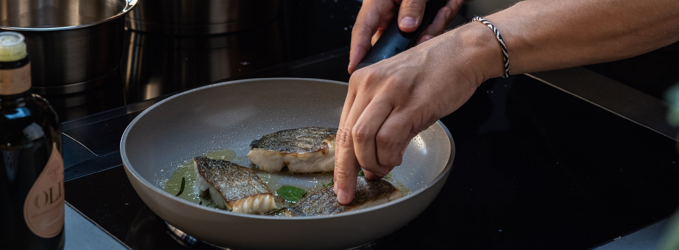 Die Hände braten fachmännisch drei Filets in einer Fissler-Pfanne auf dem Herd; Olivenöl, Kräuter und unsere ikonische Flasche sind in der Nähe.