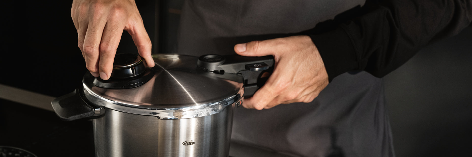 Eine Person in dunklem Hemd und grauer Schürze sichert den Deckel eines Fissler-Edelstahl-Schnellkochtopfs und stellt den Präzisionsverschluss ein.